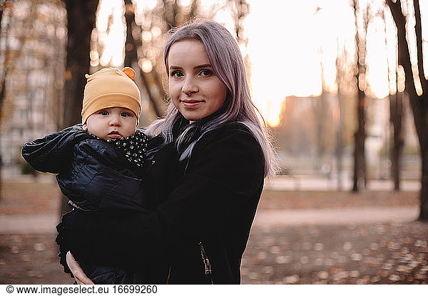 Happy young mother carrying baby son standing in park in autumn