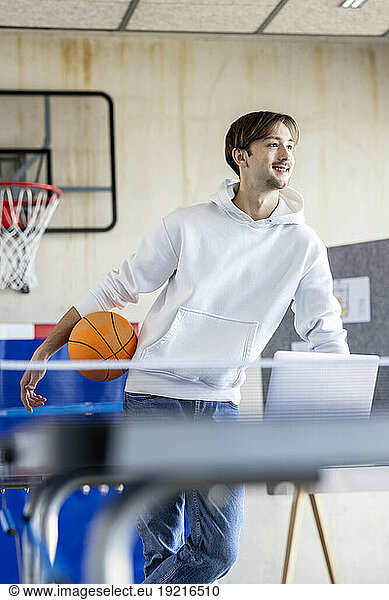 Happy young trainee standing with basketball and laptop in office