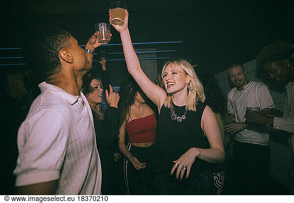 Happy young woman and man toasting drinks while dancing against friends at nightclub