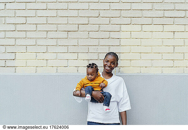Happy young woman carrying daughter standing in front of wall