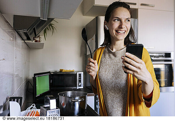 Happy young woman cooking and having video call through smart phone in kitchen
