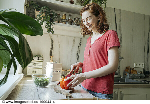 Happy young woman cutting red bell pepper in kitchen at home