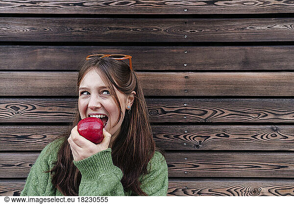 Happy young woman eating apple leaning on wooden wall