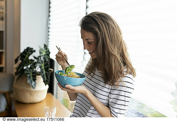 Happy young woman eating food while standing against window at home
