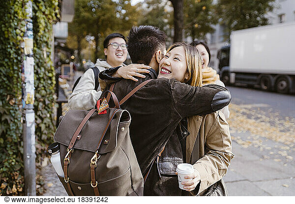 Happy young woman embracing male friend wearing backpack while standing at sidewalk