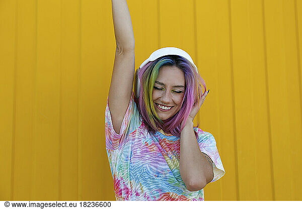Happy young woman enjoying music listening through wireless headphones and dancing in front of yellow wall