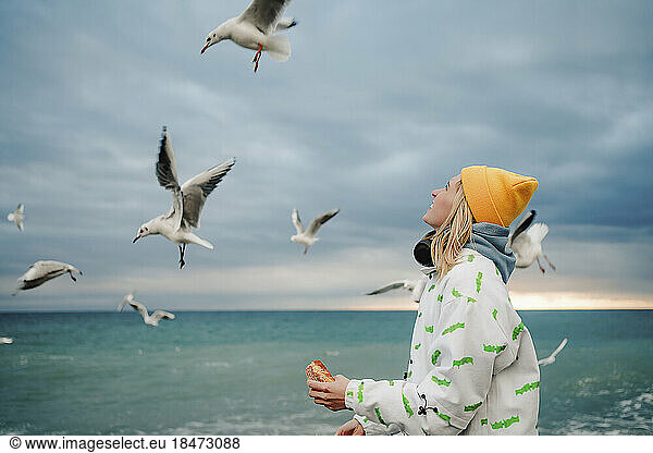 Happy young woman feeding seagulls hovering at shore
