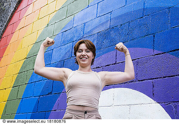 Happy young woman flexing muscles in front of rainbow flag painted on wall