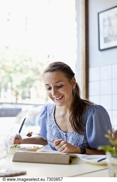 Happy young woman having meal while using digital tablet at restaurant table