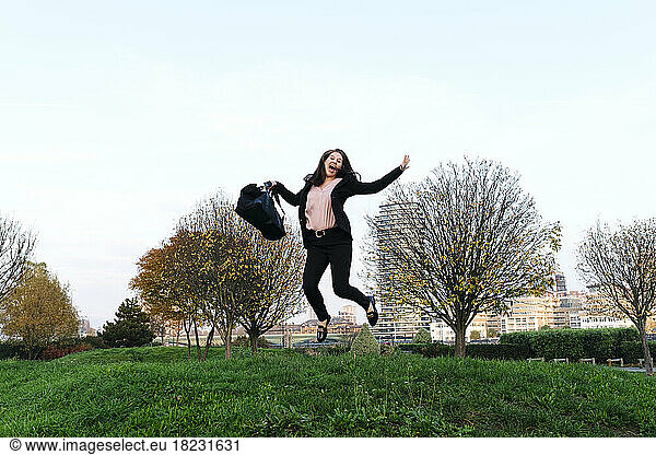 Happy young woman holding bag jumping in park