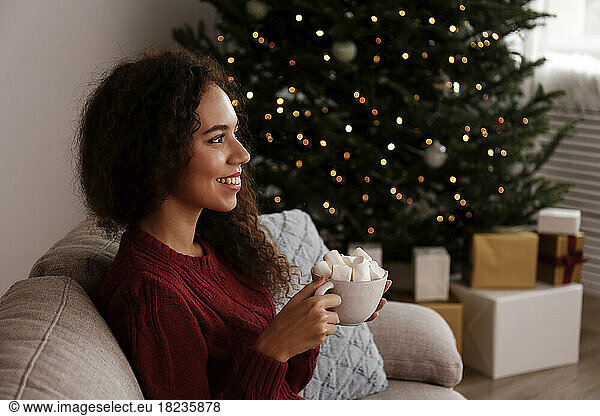 Happy young woman holding cup of marshmallow cocoa sitting on sofa at home