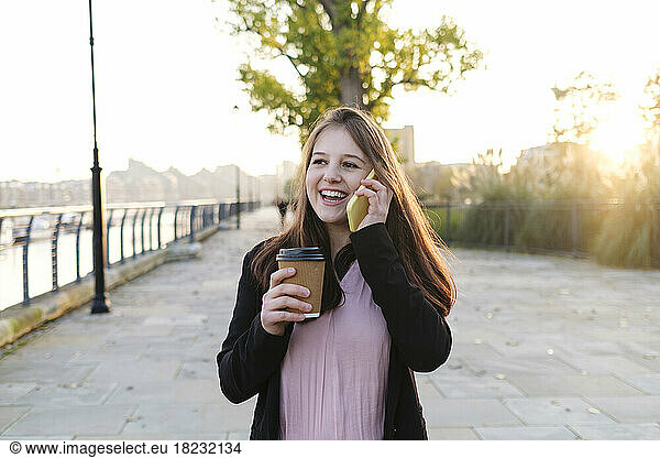 Happy young woman holding disposable cup talking on phone