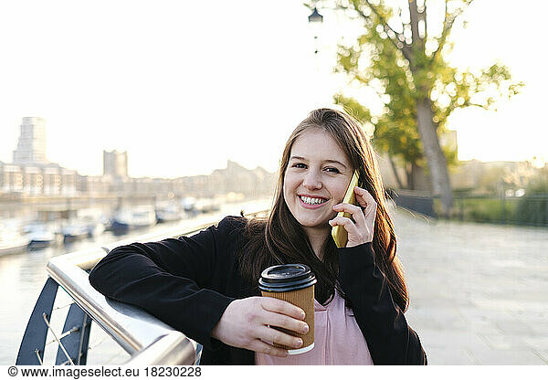 Happy young woman holding disposable cup talking on phone by railing