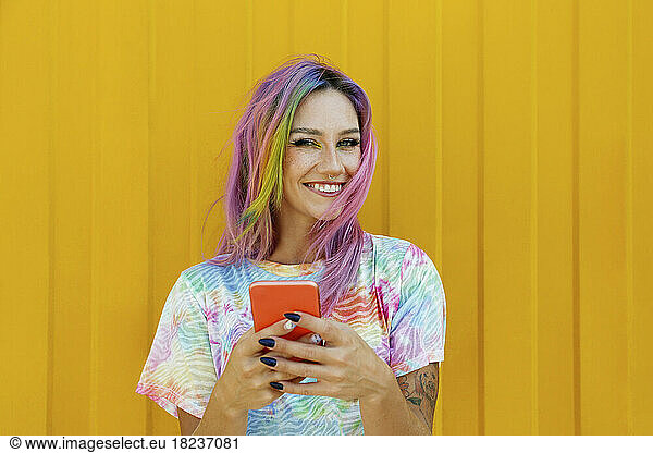 Happy young woman holding mobile phone in front of yellow wall