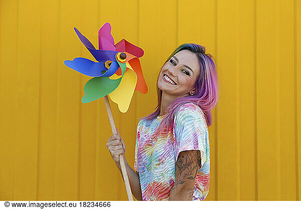 Happy young woman holding multi colored pinwheel toy in front of yellow wall