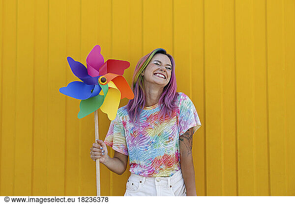 Happy young woman holding rainbow colored pinwheel toy enjoying in front of yellow wall