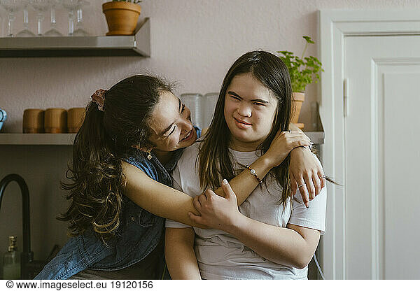 Happy young woman hugging sister with down syndrome in kitchen at home