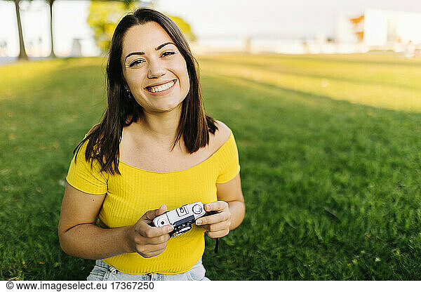 Happy young woman in off shoulder top holding camera at lawn