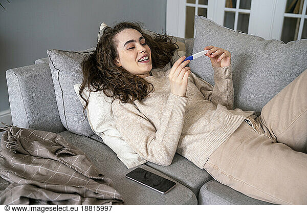 Happy young woman looking at pregnancy testing kit relaxing on sofa