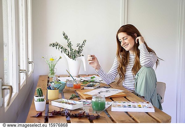 Happy young woman painting with watercolor paints on table at home