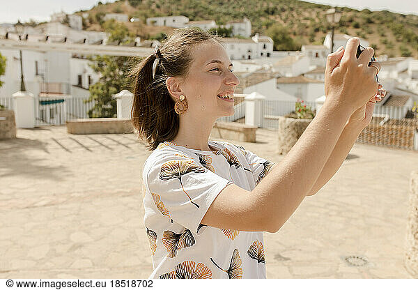 Happy young woman photographing through mobile phone on sunny day