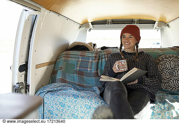 Happy young woman relaxing with book and tea in camper van