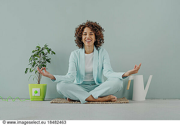 Happy young woman sitting by houseplant and watering can in front of blue background