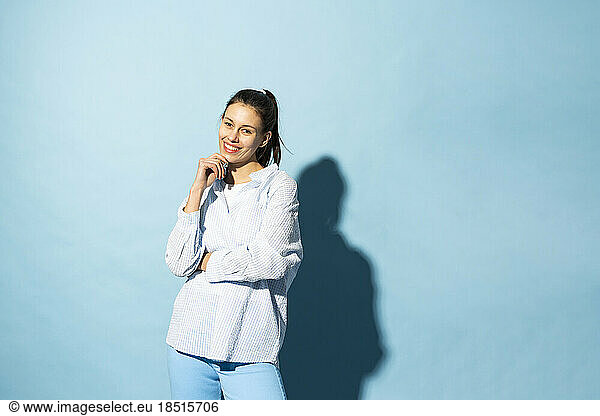 Happy young woman standing with hand on chin against blue background