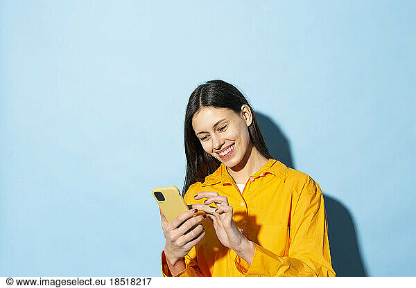 Happy young woman text messaging using smart phone against blue background