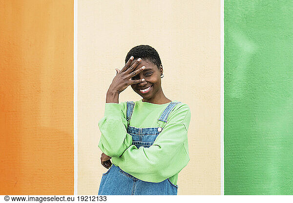 Happy young woman touching forehead standing in front of multi colored wall