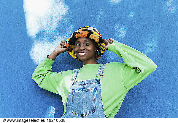 Happy young woman touching hat standing in front of blue wall