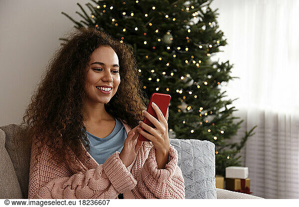 Happy young woman using smart phone in front of Christmas tree at home