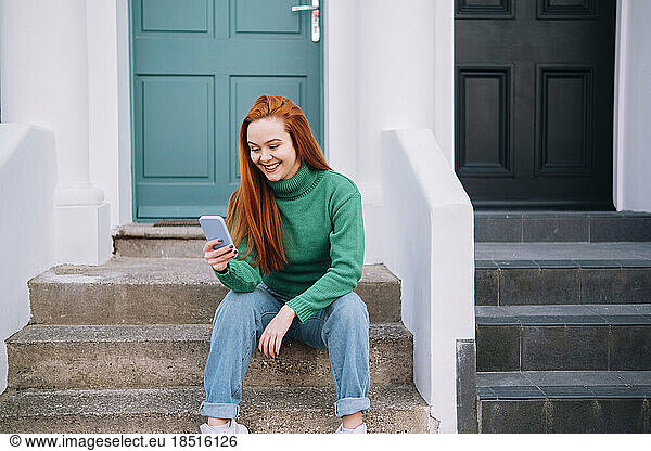 Happy young woman using smart phone sitting on steps outside house