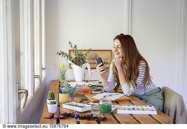 Happy young woman using smart phone while painting on table at home