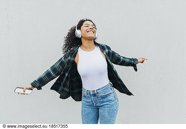 Happy young woman wearing wireless headphones dancing in front of wall