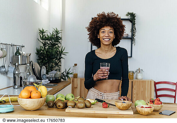 Happy young woman with Afro hairstyle holding glass of smoothie standing at table in kitchen