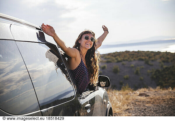 Happy young woman with arms raised leaning out of car window