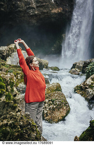 Happy young woman with arms raised standing in front of waterfall