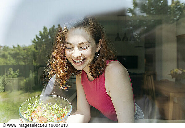 Happy young woman with bowl of salad seen through glass