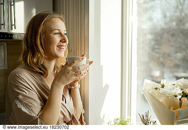 Happy young woman with coffee cup looking out through window
