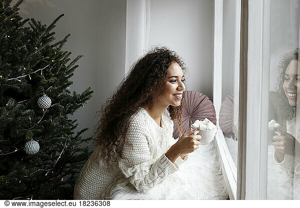 Happy young woman with cup of marshmallow cocoa looking out through window