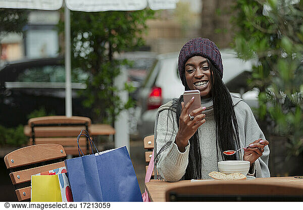Happy young woman with smart phone eating soup at sidewalk cafe