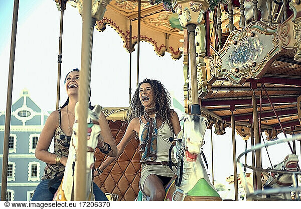 Happy young women laughing while riding carousel horses in amusement park