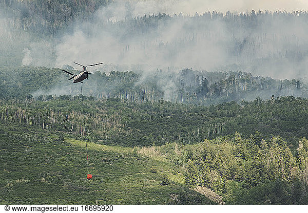 Helicopter flying with fire retardant while smoke emitting from wildfire in forest