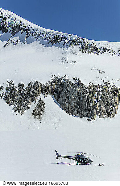 Helicopter parked on snow covered glacier below rocky mountain ridge.