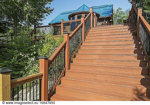 Hellbraun gebeizte Holztreppe mit schwarzem schmiedeeisernem Geländer  die zu einem braun gebeizten  gefrästen östlichen Weißkiefernholz und einem flachen Blockhaus mit blauem Stehfalzblechdach im Sommer in Quebec  Kanada  hinaufführt. Dieses Bild ist als Eigentum freigegeben. CUPR0362.