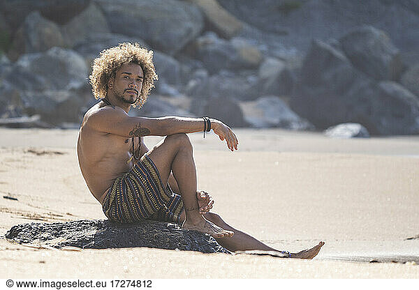 Hemdloser junger Mann sitzt auf einem Felsen am Strand im Sommerurlaub