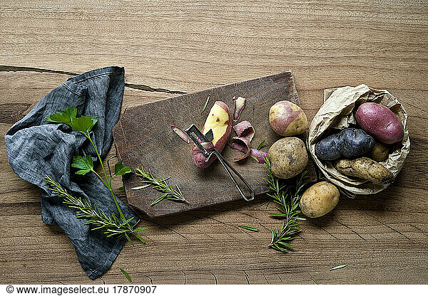 Herbs  dish towel  cutting board  potato peeler and different varieties of raw potatoes