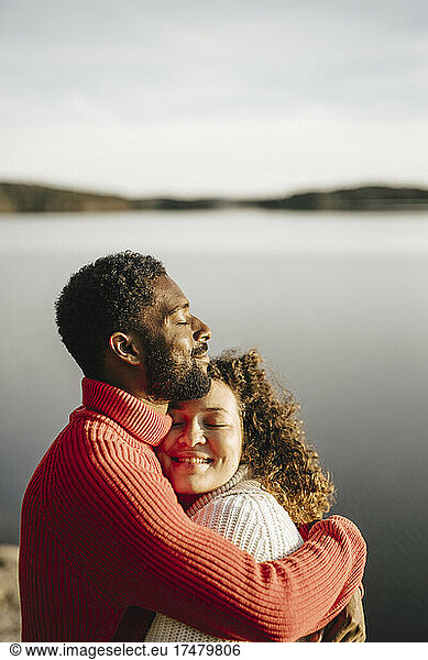 Heterosexual couple embracing by lake on sunny day