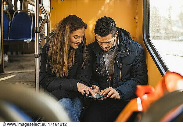 Heterosexual couple using smart phone while sitting in tram during pandemic